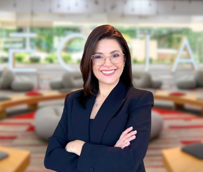 Una mujer sonriente con gafas y traje negro está posando en un entorno moderno y luminoso.