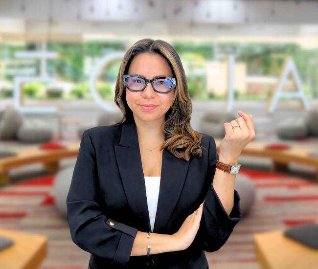 Una mujer con gafas y traje oscuro sonríe frente a un fondo moderno.