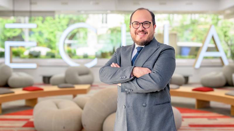 Un hombre sonriente con gafas y un traje gris posando en un ambiente moderno y acogedor.