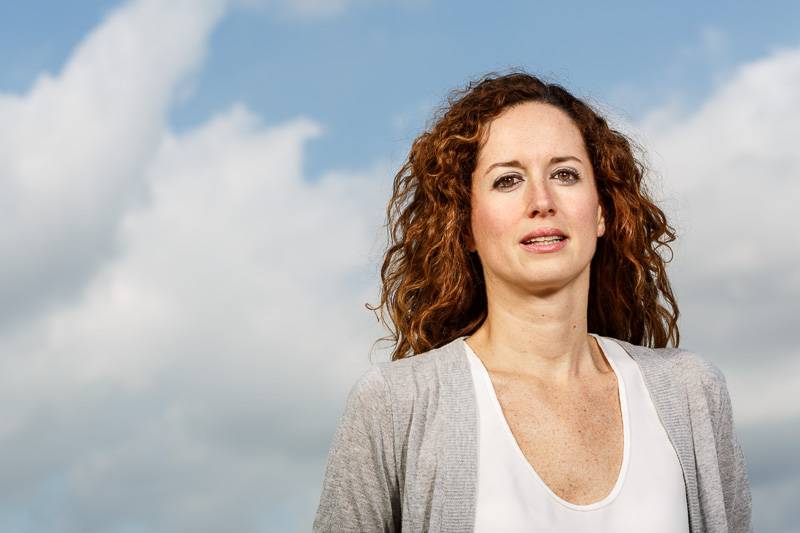Mujer con cabello rizado y prendas claras posa frente a un cielo nuboso.