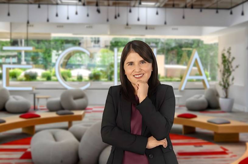 Una mujer sonriente se encuentra en un ambiente moderno y acogedor, con muebles contemporáneos y plantas decorativas de fondo.