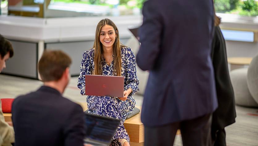 Una joven sonriente está sentada con una computadora portátil en un ambiente moderno y luminoso.
