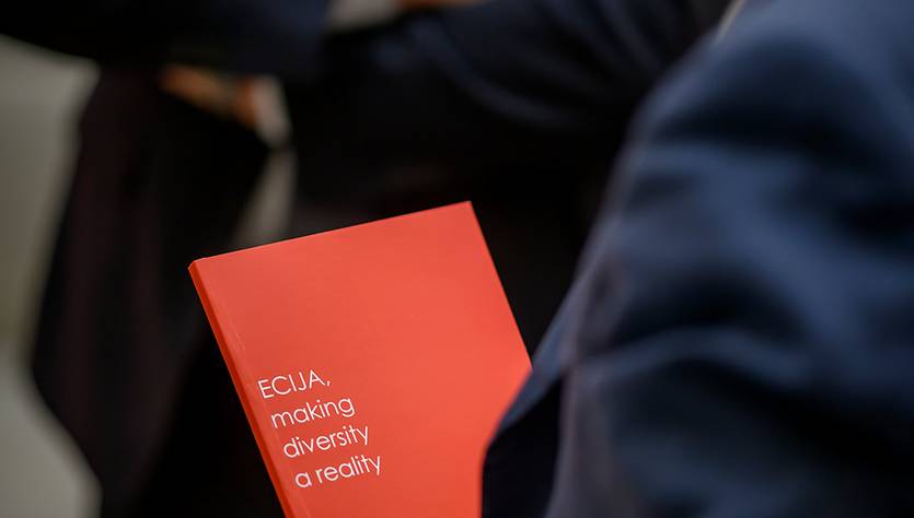 Un hombre sostiene un documento de color rojo que dice 'ECLJA: making diversity a reality', mientras dos personas están al fondo.