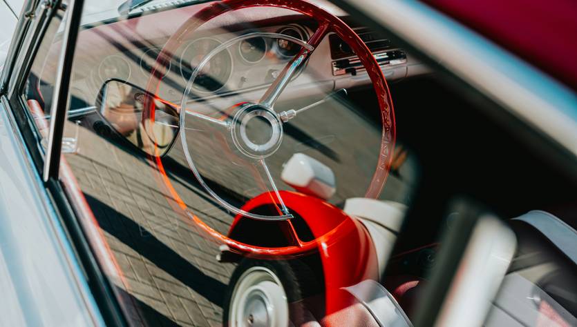 La imagen muestra el interior de un automóvil clásico con un volante de color rojo y asientos en tonos claros.