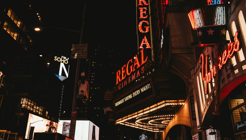 La imagen muestra un cine Regal iluminado por neón en una concurrida calle de la ciudad durante la noche.