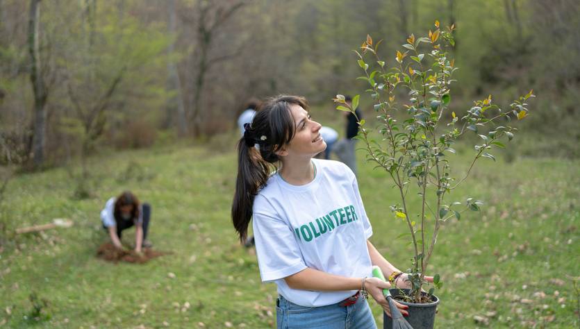 Una mujer sonriente sostiene una planta en un entorno natural mientras otros voluntarios trabajan en el fondo.