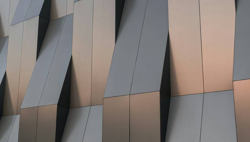 La imagen muestra una fachada arquitectónica con paneles metálicos en forma ondulada y tonos grises y dorados.