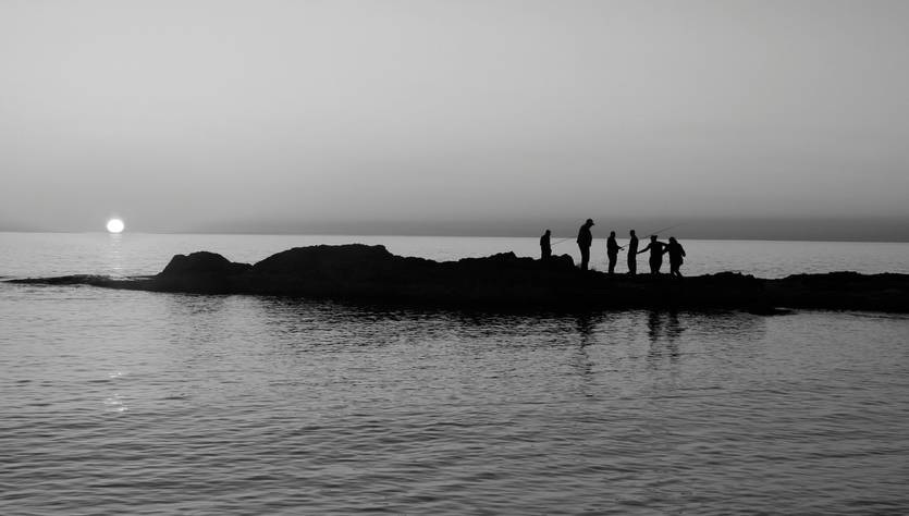 Siluetas de personas en una roca al atardecer, con el sol poniéndose en el horizonte.