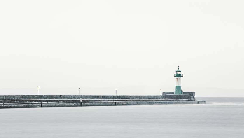 Un faro solitario se alza en un muelle rodeado por un mar tranquilo en un día nublado.