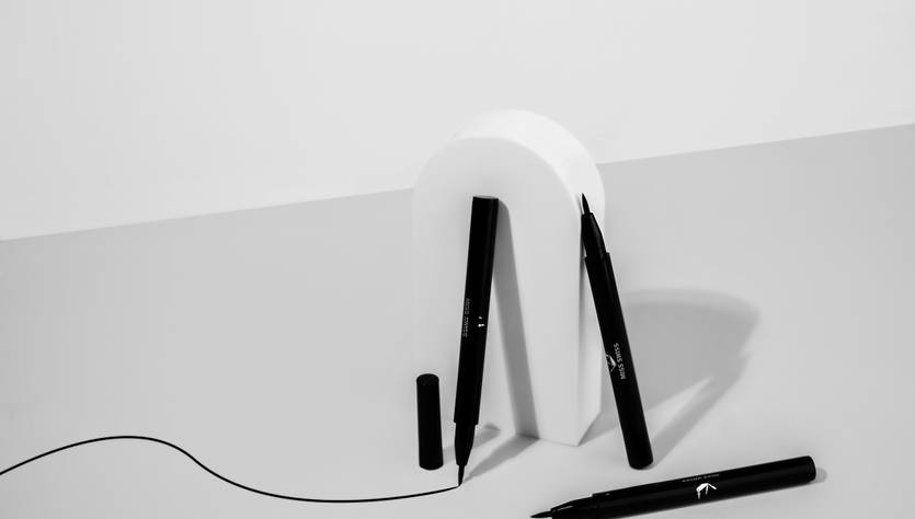 Una composición artística que presenta un soporte blanco en forma de letra y varios marcadores negros en una mesa clara.