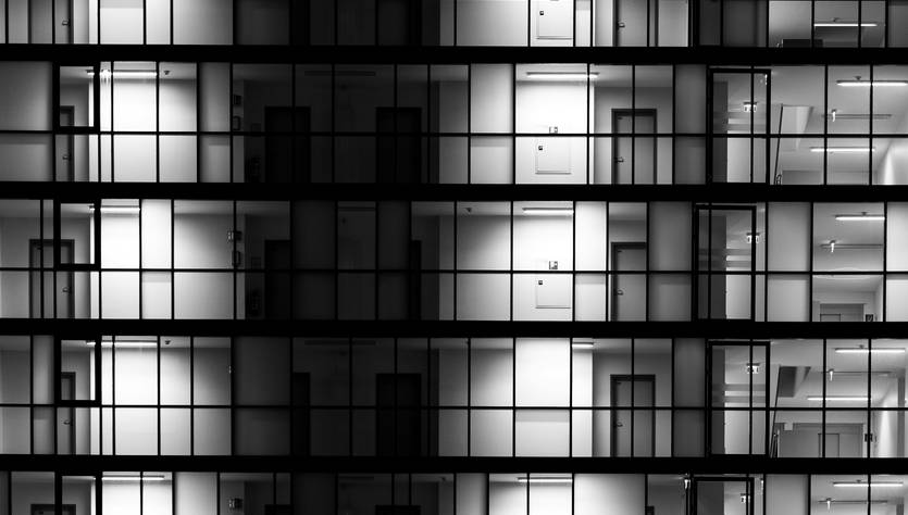 La imagen muestra una fachada de un edificio iluminado en blanco y negro, destacando filas de ventanas con distintos niveles de luz.
