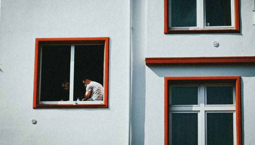 Una persona observa desde una ventana con marcos rojos en un edificio blanco.