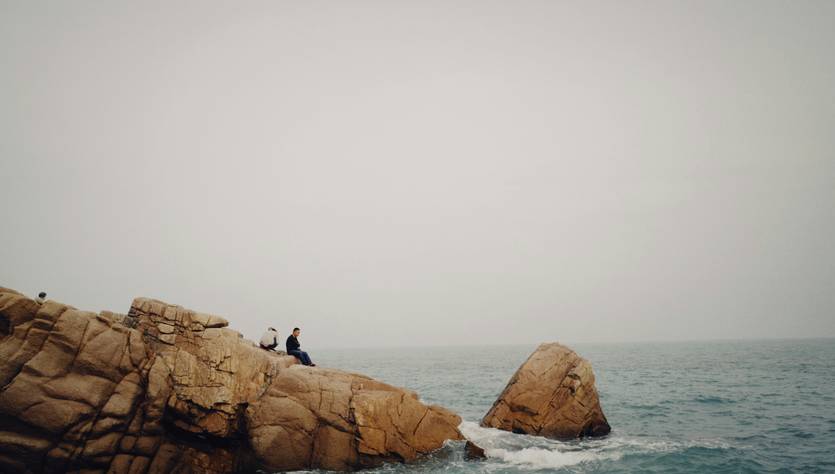 Dos personas sentadas en una roca frente al mar en un día nublado.