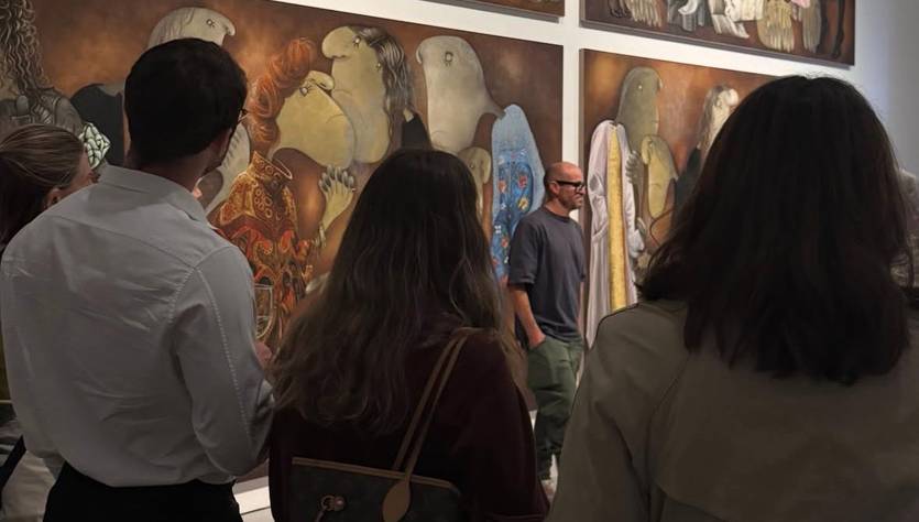 Un grupo de personas observa una exposición de arte en una galería, junto a grandes obras que representan figuras estilizadas.