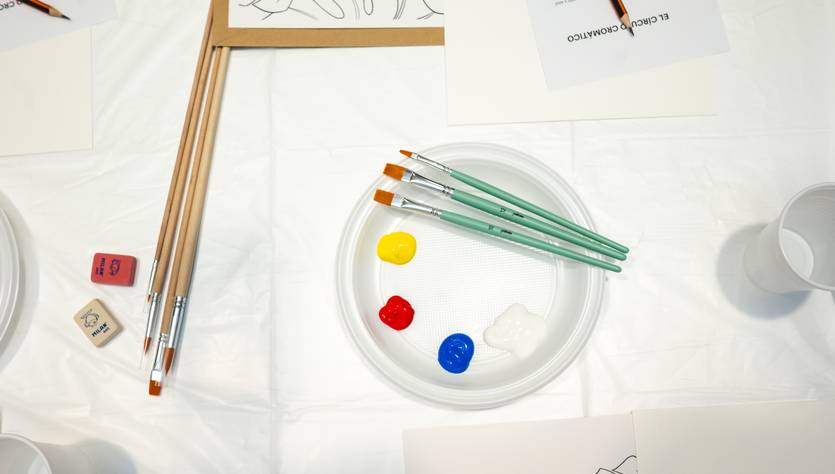 La imagen muestra una mesa con pinceles, pinturas y hojas para colorear preparadas para una actividad artística.