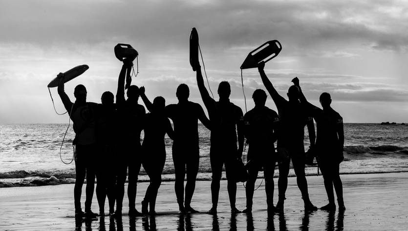 Un grupo de salvavidas se reúne en la playa al atardecer, levantando sus salvavidas en señal de unidad.