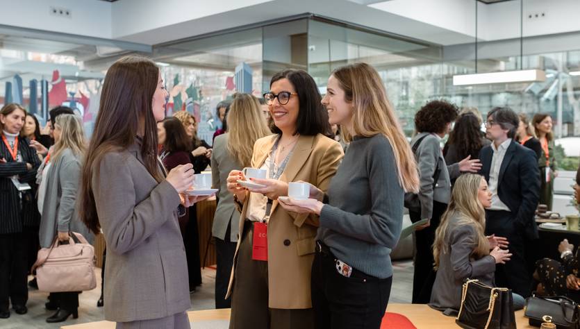 Tres mujeres conversan y comparten una bebida en un evento social rodeadas de otras personas.