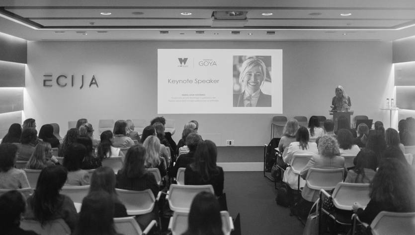 La imagen muestra a un grupo de personas sentadas en un evento, escuchando a una ponente que se presenta en pantalla.