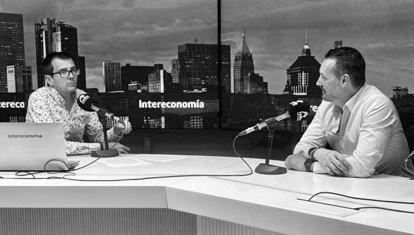 Dos hombres conversan en un estudio de televisión con un fondo urbano.