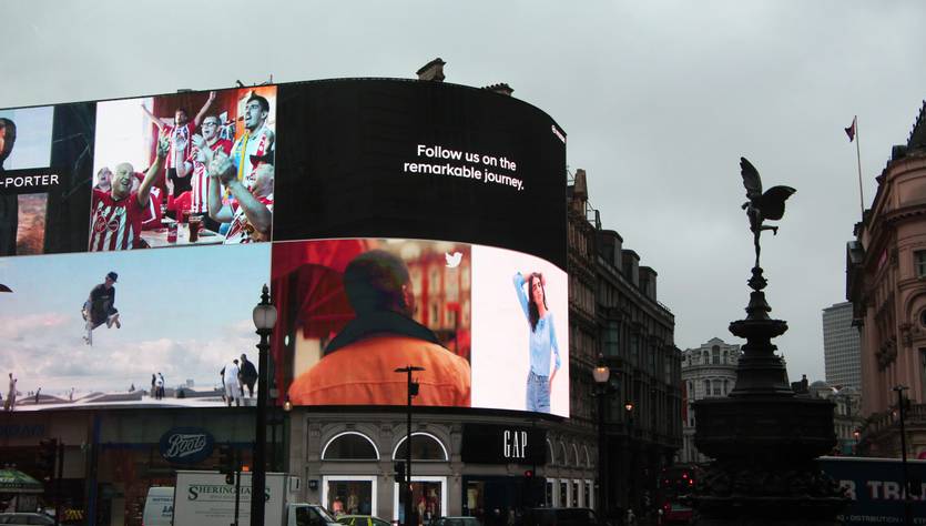 Una imagen de un cartel publicitario iluminado en una plaza de la ciudad con diversas imágenes y un mensaje inspirador.