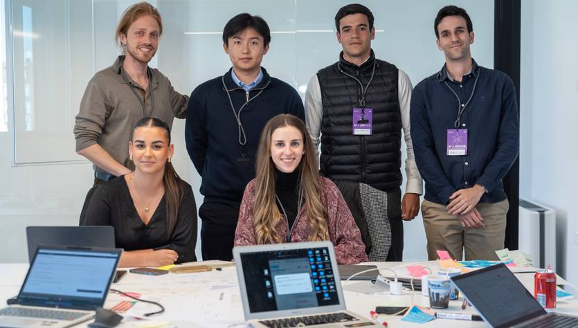 Un grupo de seis personas posando juntas en una oficina moderna con computadoras portátiles y documentos en una mesa.