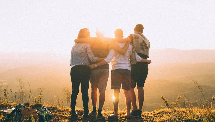Un grupo de cuatro personas se abraza frente a un paisaje iluminado por el sol al atardecer.