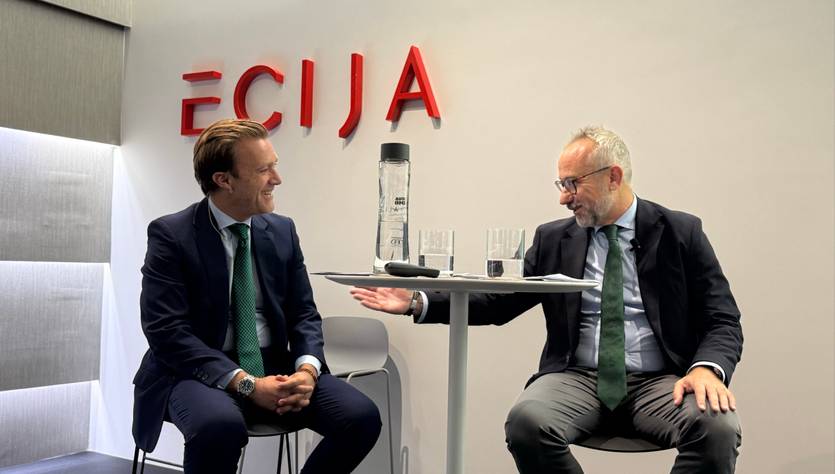 Dos hombres en una conversación en un ambiente profesional bajo un letrero con el nombre 'ECIJA'.