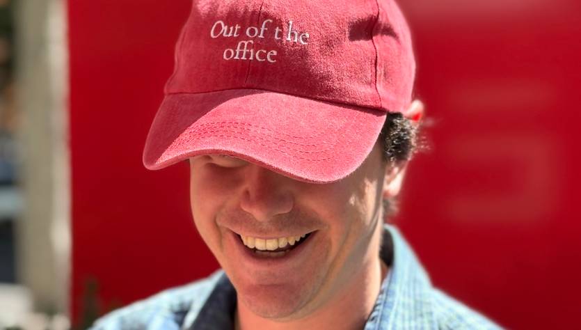 Un hombre sonriente lleva una gorra roja con la frase 'Out of the office'.