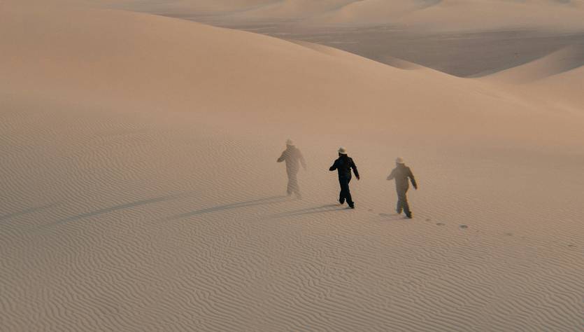 Tres personas caminan por un desierto de arena en condiciones de viento.