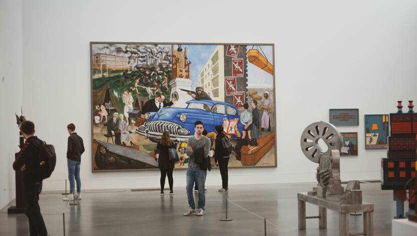 Una sala de museo con visitantes observando una gran pintura de un automóvil azul y diversas figuras humanas y elementos arquitectónicos en un entorno urbano.