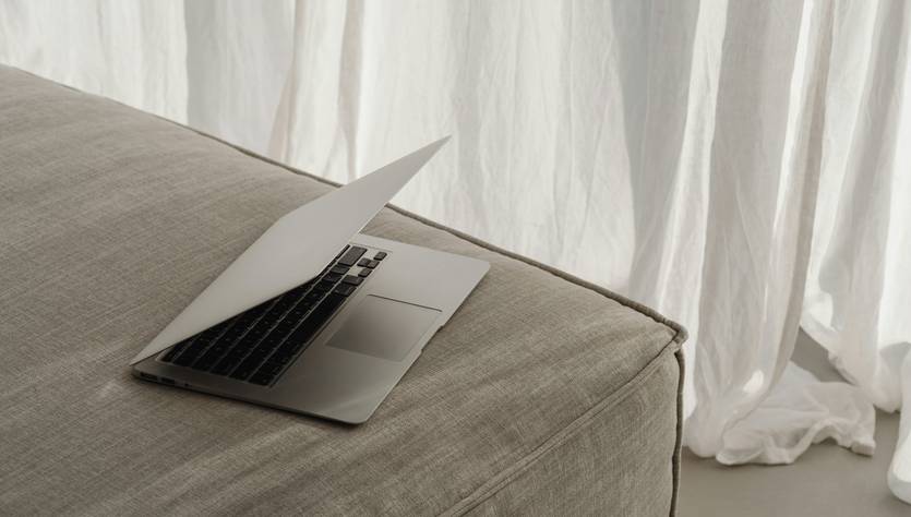 Una laptop abierta sobre un sofá de tela clara junto a una cortina blanca.