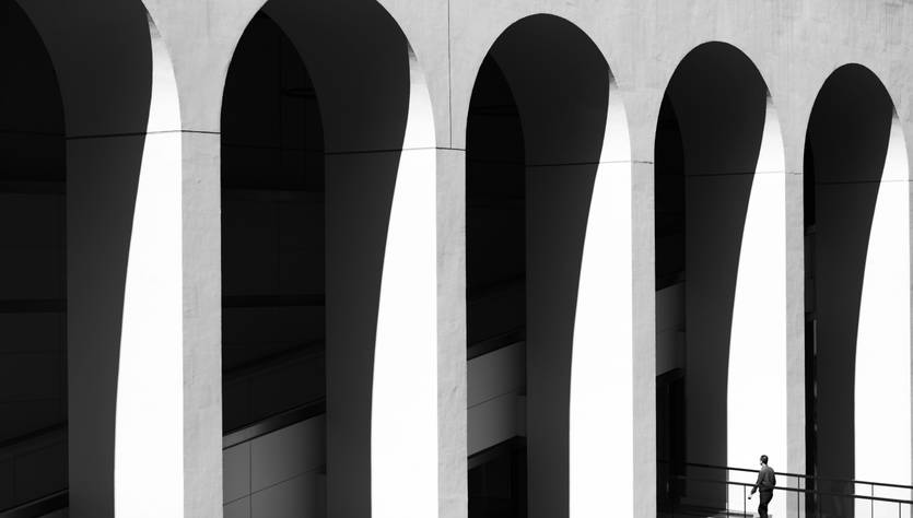 Una persona camina bajo arcos en un entorno arquitectónico en blanco y negro.