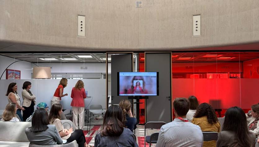 Un grupo de personas se reúne en una sala moderna con una pantalla que muestra a otra persona en videoconferencia.