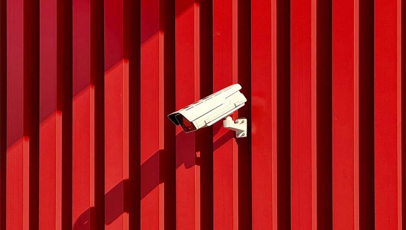 La imagen muestra una cámara de seguridad montada sobre una pared roja con líneas verticales.