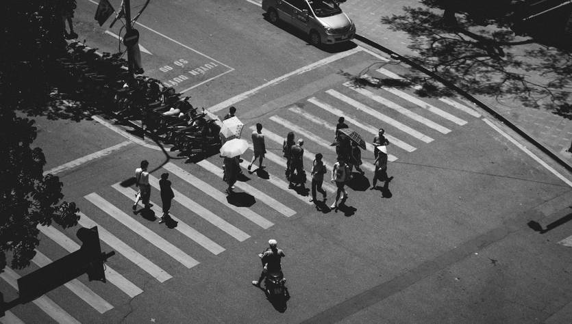 Una imagen en blanco y negro que muestra un grupo de personas cruzando una calle con un paraguas en una zona urbana.