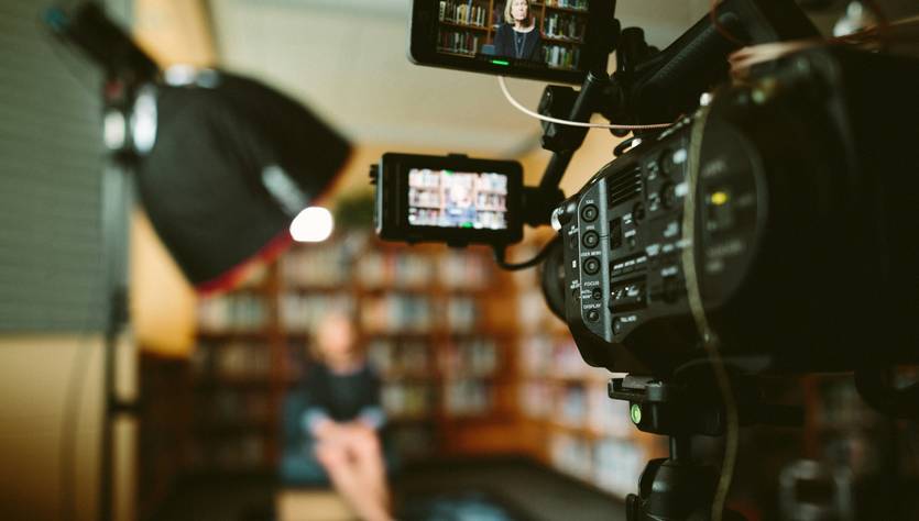 Una cámara de video en primer plano enfocando a una persona sentada en un entorno de estudio con estanterías de libros de fondo.