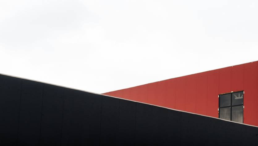 La imagen muestra una composición arquitectónica con estructuras de colores negro y rojo bajo un cielo nublado.