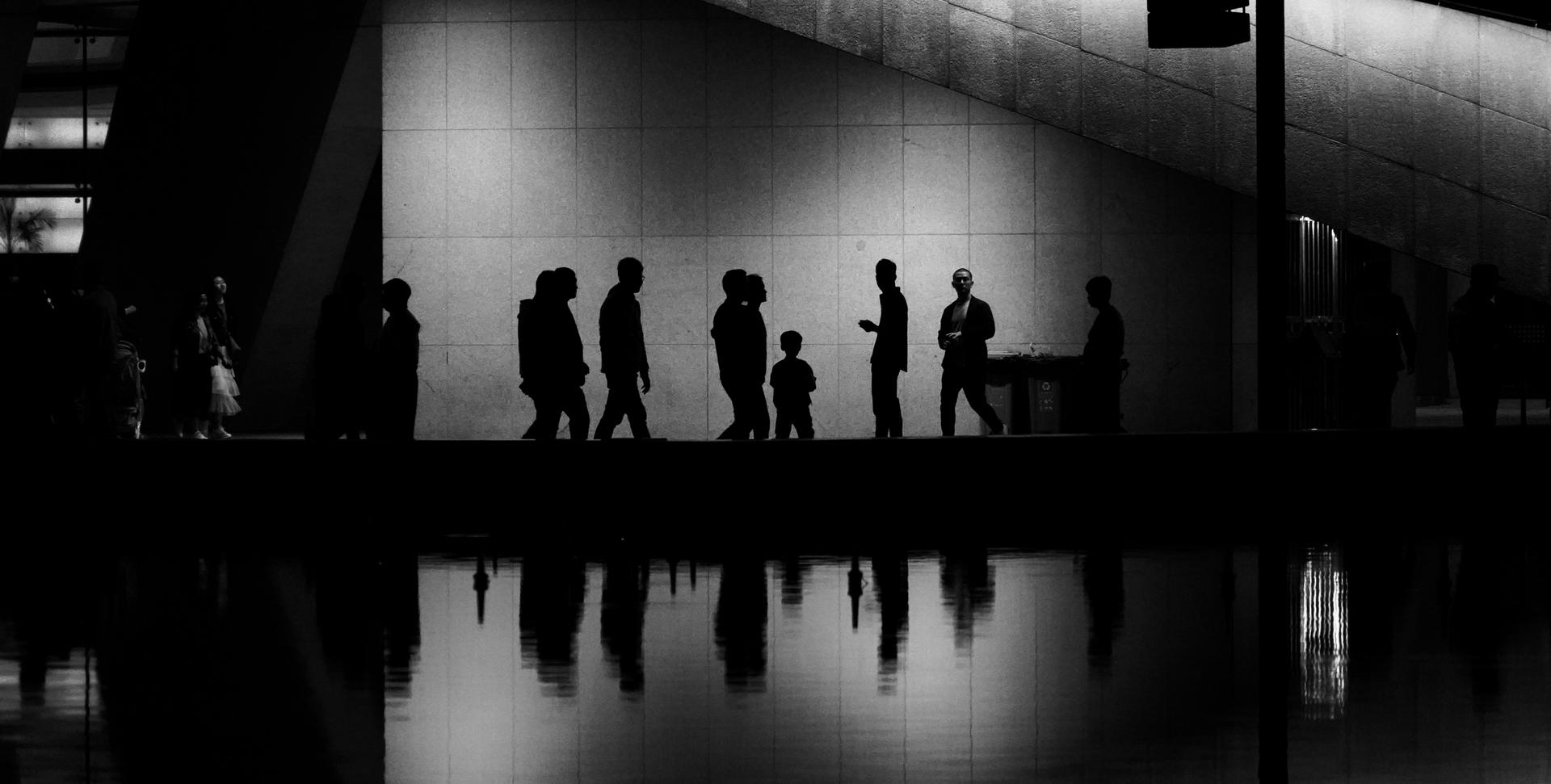 Siluetas de personas caminando a lo largo de un muro iluminado, reflejándose en el agua.