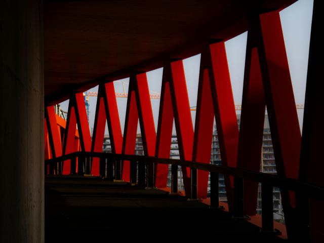 Una estructura arquitectónica con soportes rojos y un pasillo oscuro que muestra edificios en construcción al fondo.