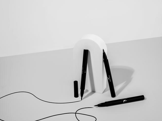 Una composición artística que presenta un soporte blanco en forma de letra y varios marcadores negros en una mesa clara.