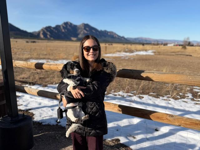 Una joven sostiene un perro en un paisaje montañoso con cielo despejado.