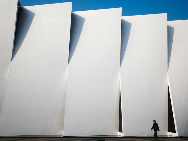 Una persona camina frente a una moderna y abstracta fachada blanca con ángulos sobresalientes.