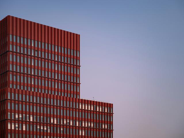 Un edificio moderno de color rojo con ventanales, destacándose en un cielo suave al atardecer.