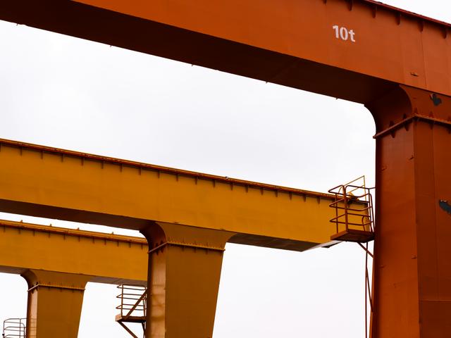 La imagen muestra grúas de carga de color naranja y amarillo con un cielo gris al fondo.
