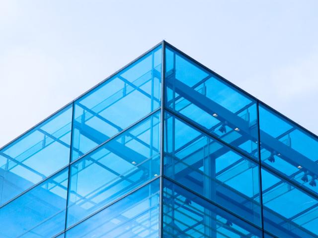 La imagen muestra la esquina superior de un edificio moderno con una fachada de cristal azul.