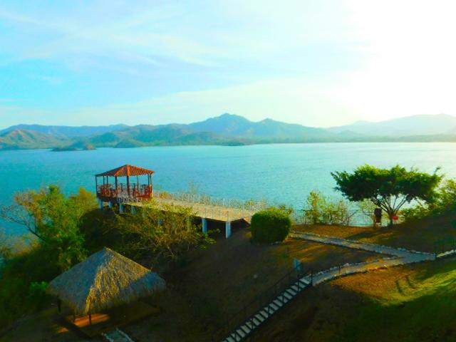 Vista panorámica de un lago rodeado de montañas y vegetación, con un muelle y una construcción a la orilla.
