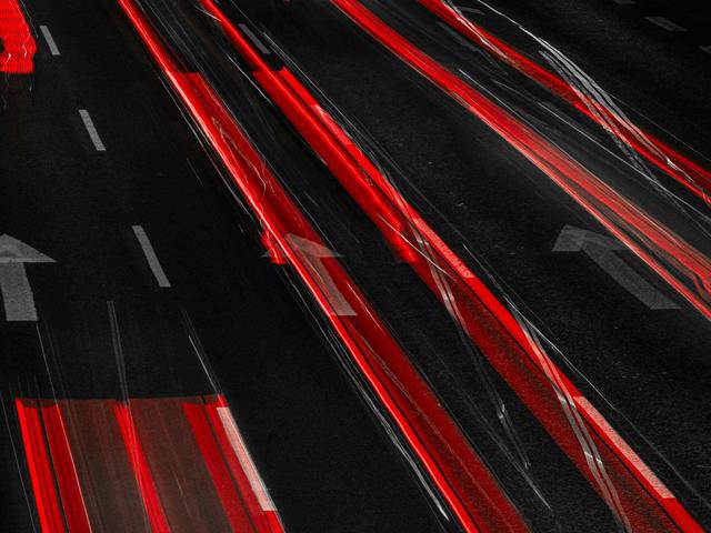 La imagen muestra el movimiento de vehículos en una carretera oscura, con luces rojas dejando estelas brillantes.