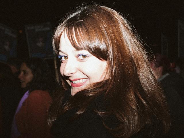 Una mujer sonriente se encuentra en un entorno de evento con personas al fondo.