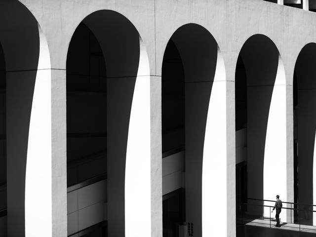 Una persona camina bajo arcos en un entorno arquitectónico en blanco y negro.