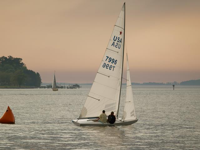 Un par de personas navegan en un velero sobre un lago al atardecer.